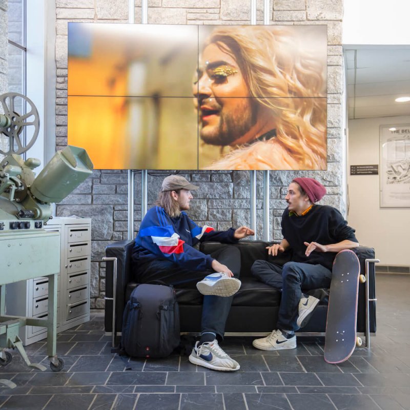 Two Falmouth University students sat on a sofa next to an old film projector and a large image of blonde bearded person behind them.
