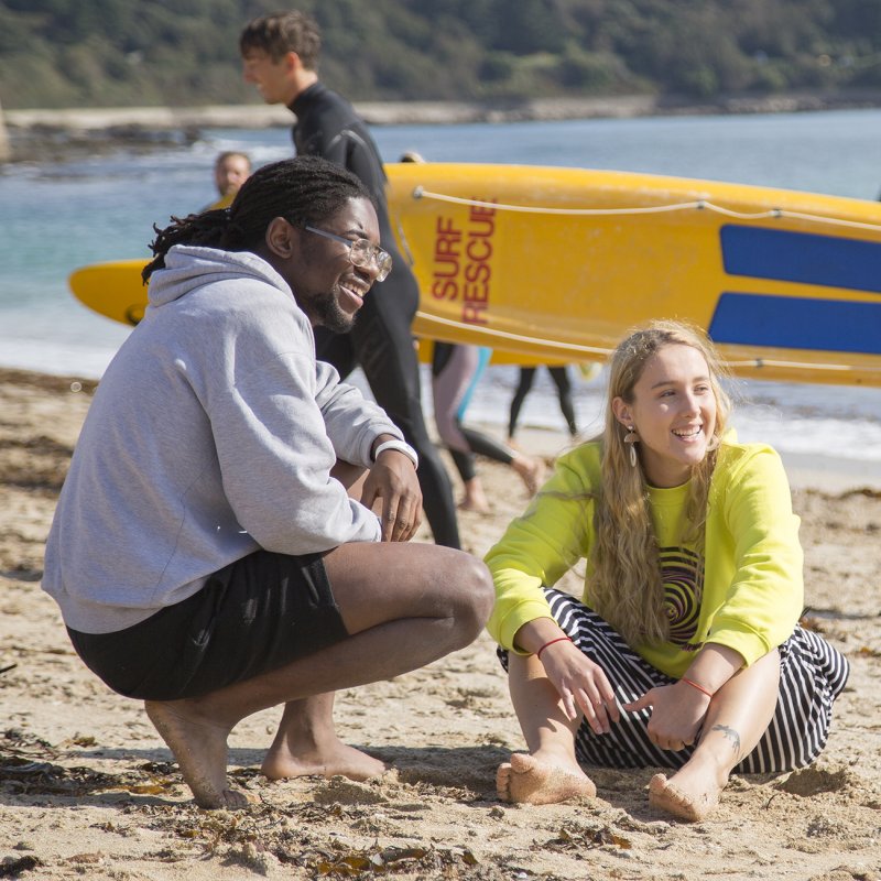 Two students sitting on the beach in front of a yellow surf rescue board
