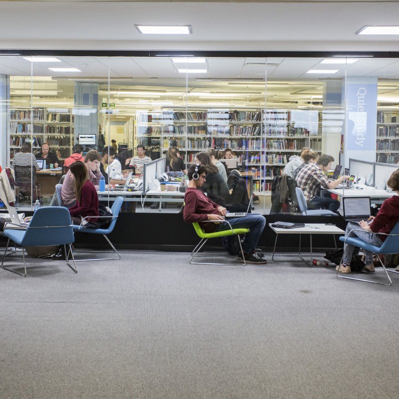 Students sat on low chairs working in front of large window looking through to the library on Penryn campus.