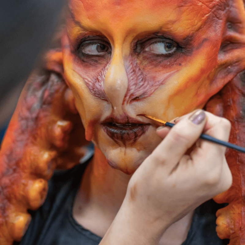 A person wearing a prosthetics orange mask with a hand and a paintbrush