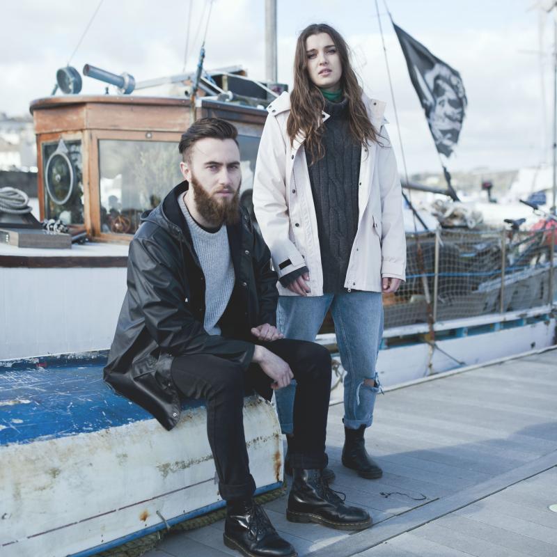 Couple posing by boat in fashionable rainwear, knitwear and black boots.