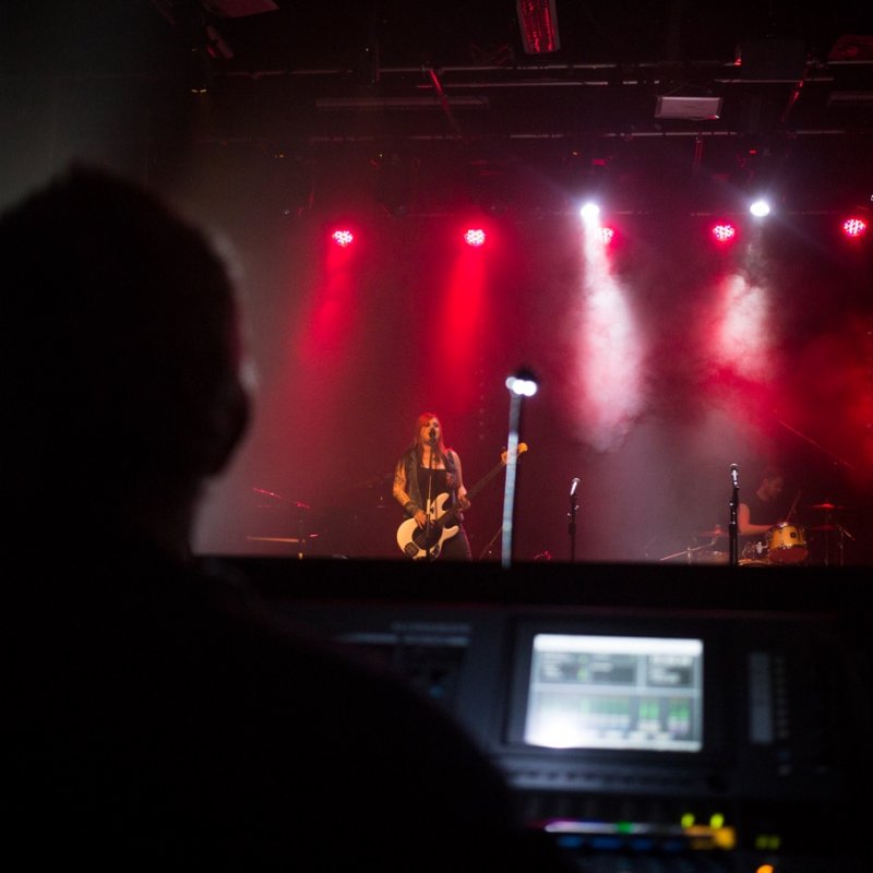 Musician playing guitar on a red lit stage, figure at mixing desk in the foreground