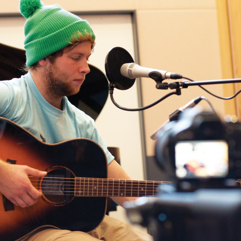 Student in green bobble hat playing guitar into microphone in studio.