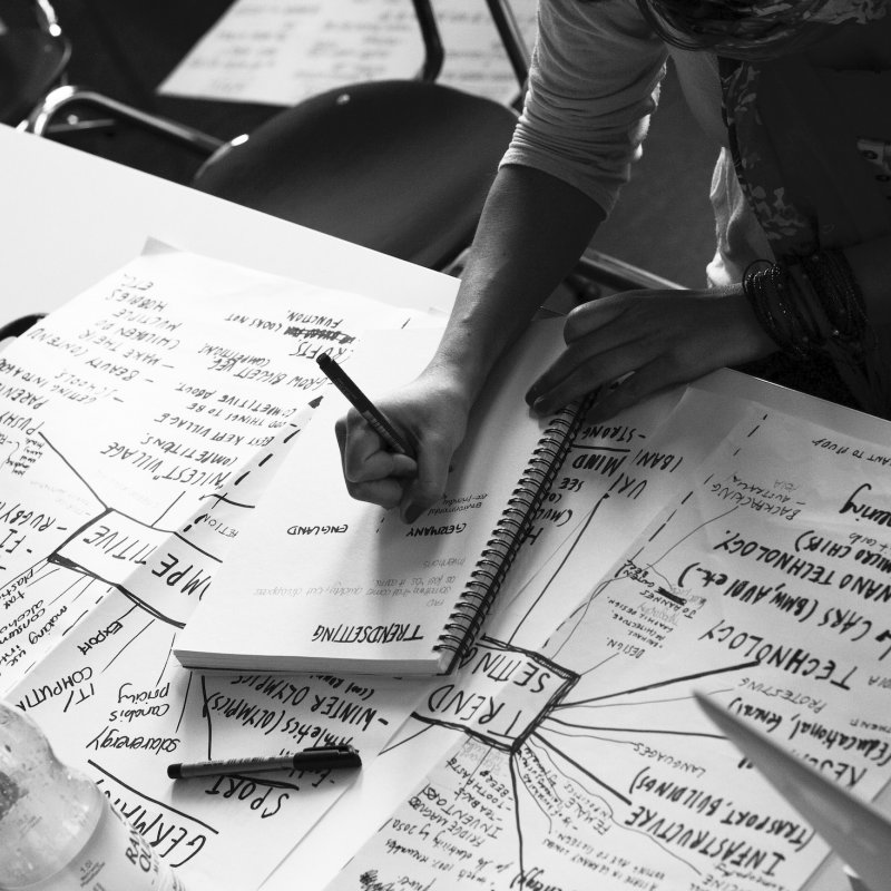 Grayscale image of students hands writing and filling pages in front of them with ideas