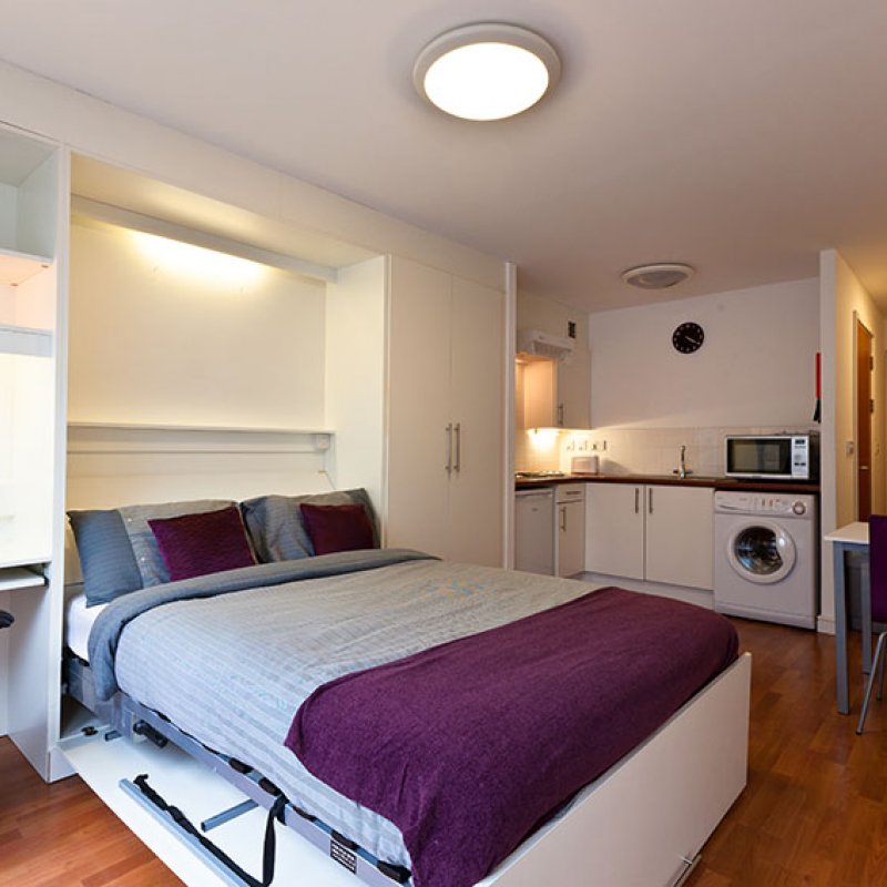 Bedroom studio interior with grey bedspread, purple cushions, and washing machine