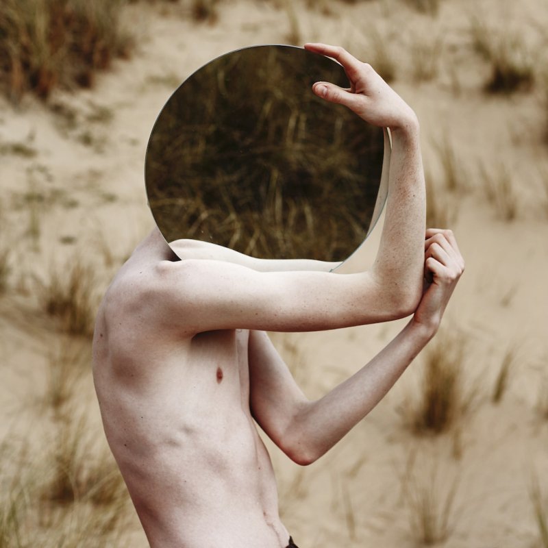 Bare chested model carrying circle mirror in front of face reflecting grass.