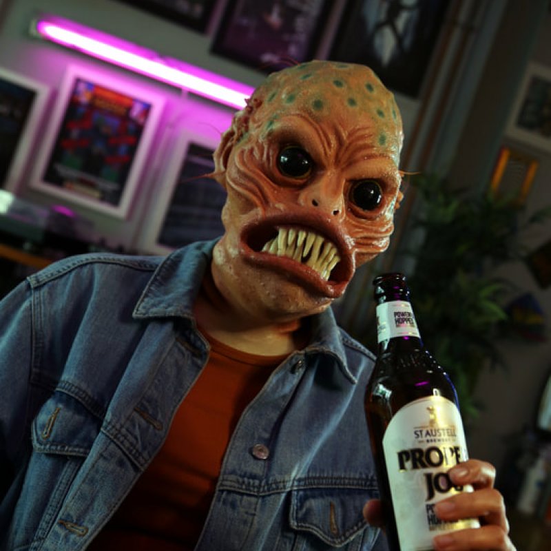 Individual wears alien prosthetic mask, holding bottle of beer