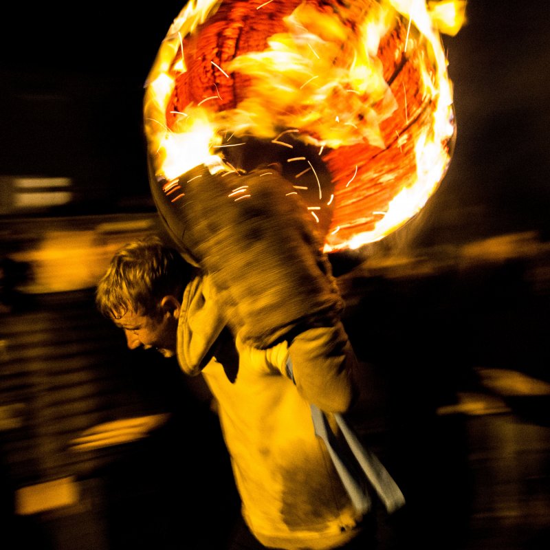 Youth carrying barrel full of fire on shoulders.