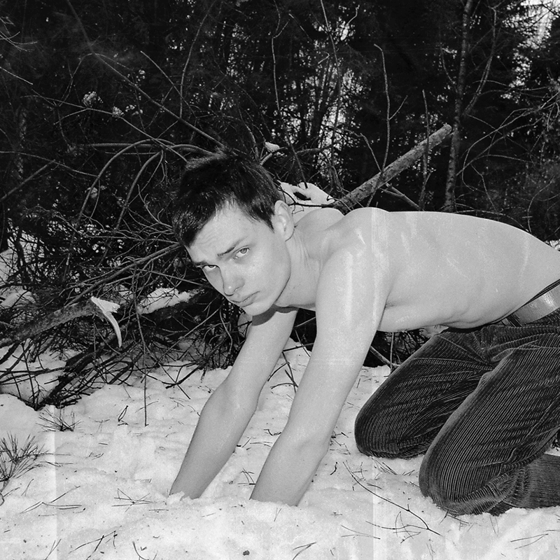 A topless man with his hands in the snow, with pine trees behind