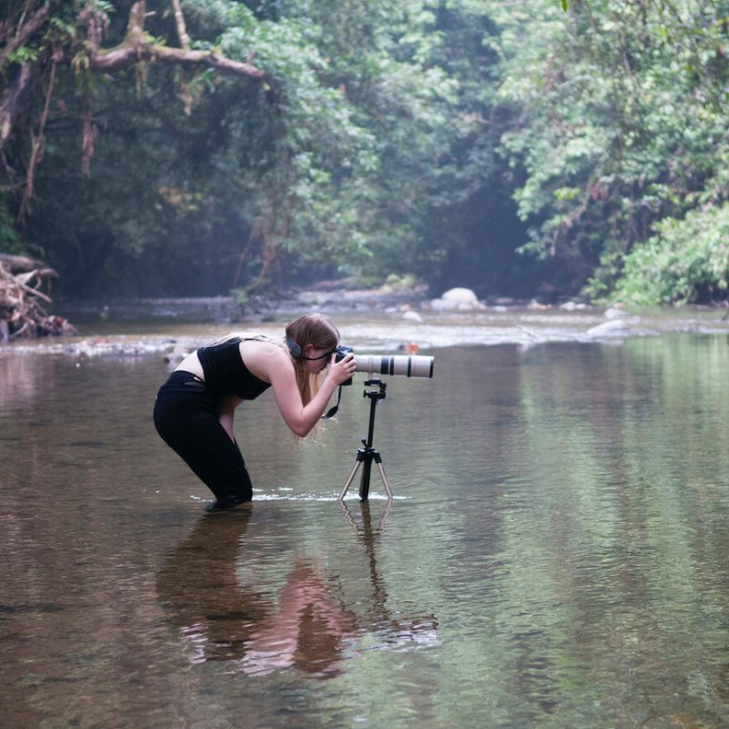 Falmouth University student immersed up to knees in river, bending down to view though camera on tripod.