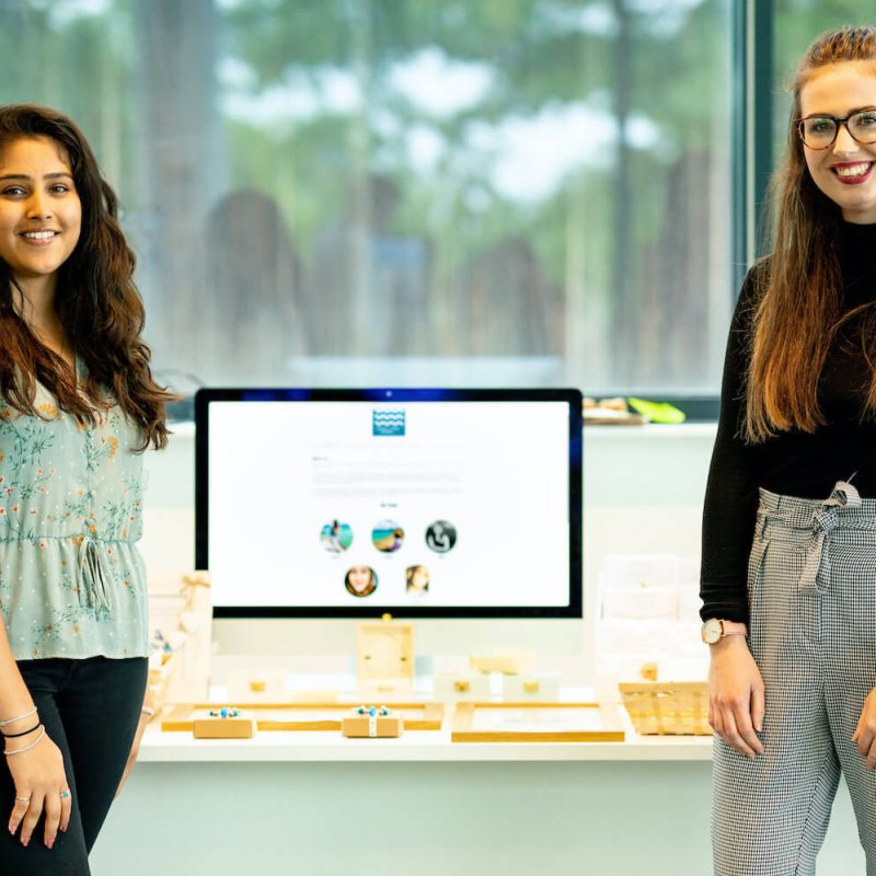 Two Falmouth University students stood between a desktop computer, smiling