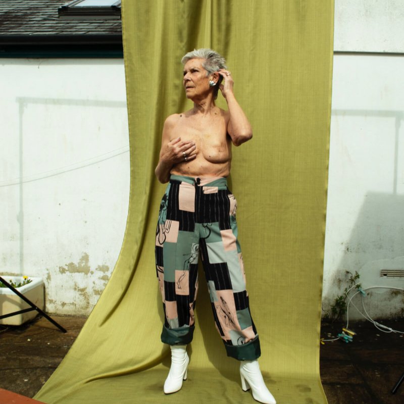 A topless woman with patchwork trousers, in front of a green curtain