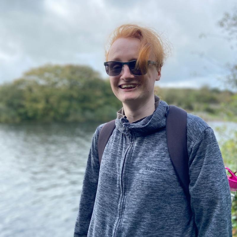 A Falmouth University students next to a water and trees, wearing a hoodie and sunglasses