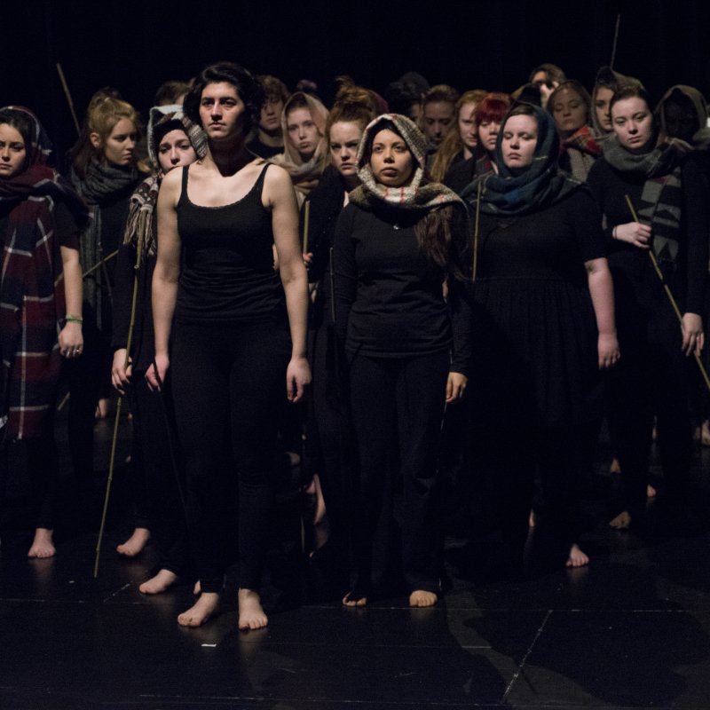 Group of students in dark clothes, head scarves and holding sticks on stage.