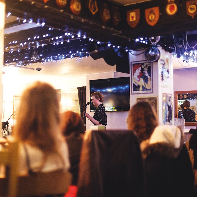 Girl reading from paper in hands in front of audience in pub.