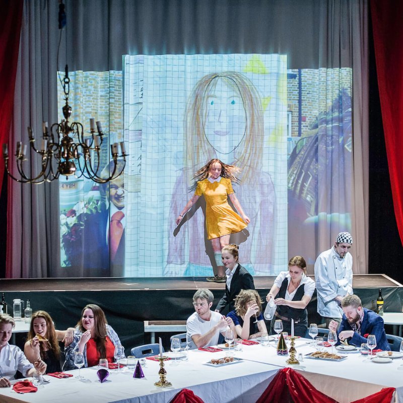 Actor in yellow dress dancing on stage in front of childlike portrait projected behind her.