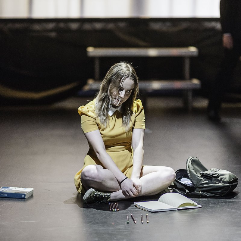 Actor in yellow dress sat cross legged on the stage.