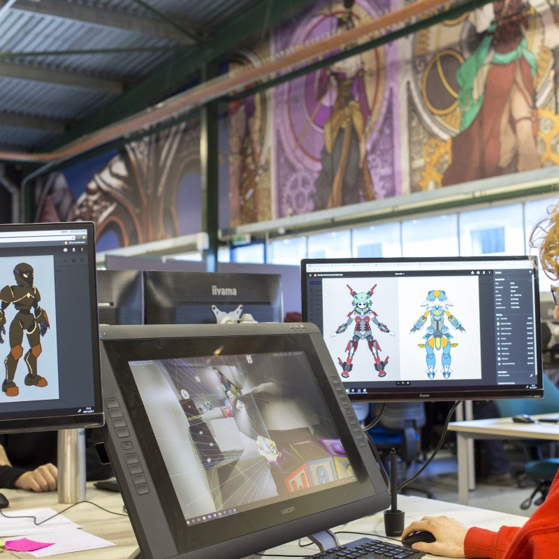 Male student at a desk with three computer screens designing digital games