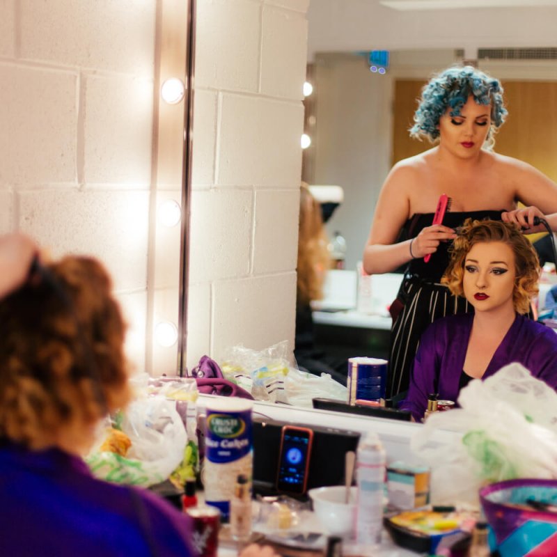 A girl getting her hair styled by another girl in a dressing room with a mirror