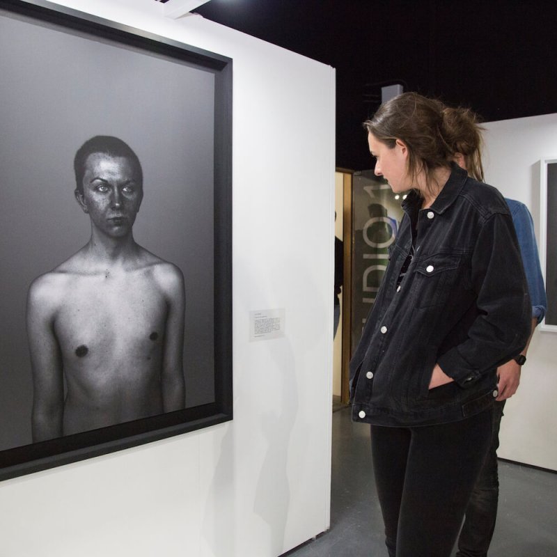 Woman looking at a framed photography portrait of a topless boy on a gallery wall.