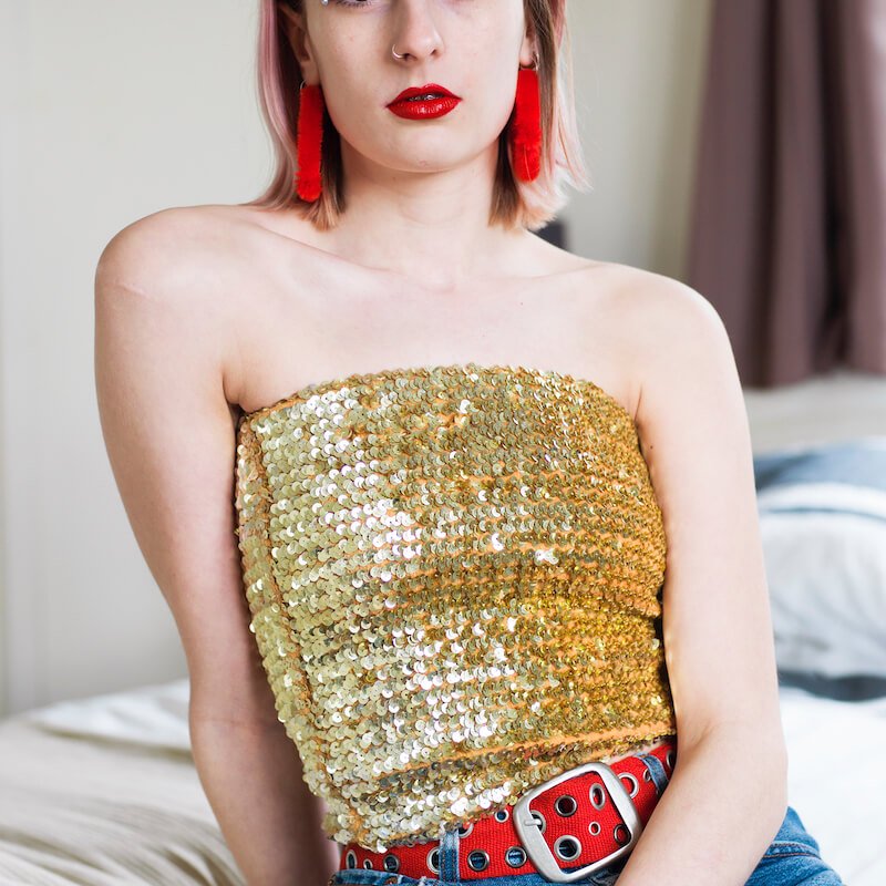 Model with gold sequin top and red tassel earring and red belt sat on a bed.