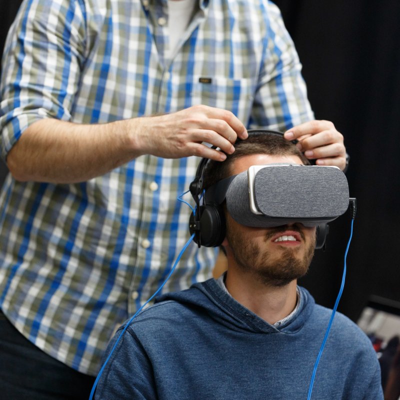 man seated wearing VR headset with another man stood behind him