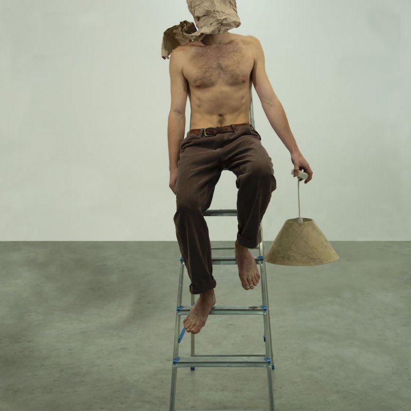 A topless man on a stool wearing brown trousers and a brown bag over his head