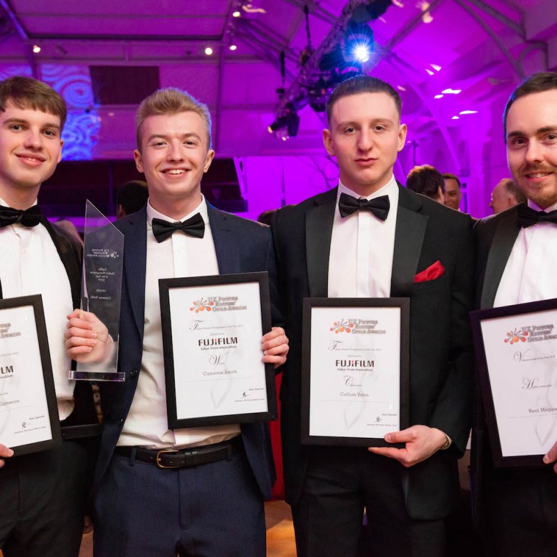 Four male students dressed in tuxedos and holding framed awards