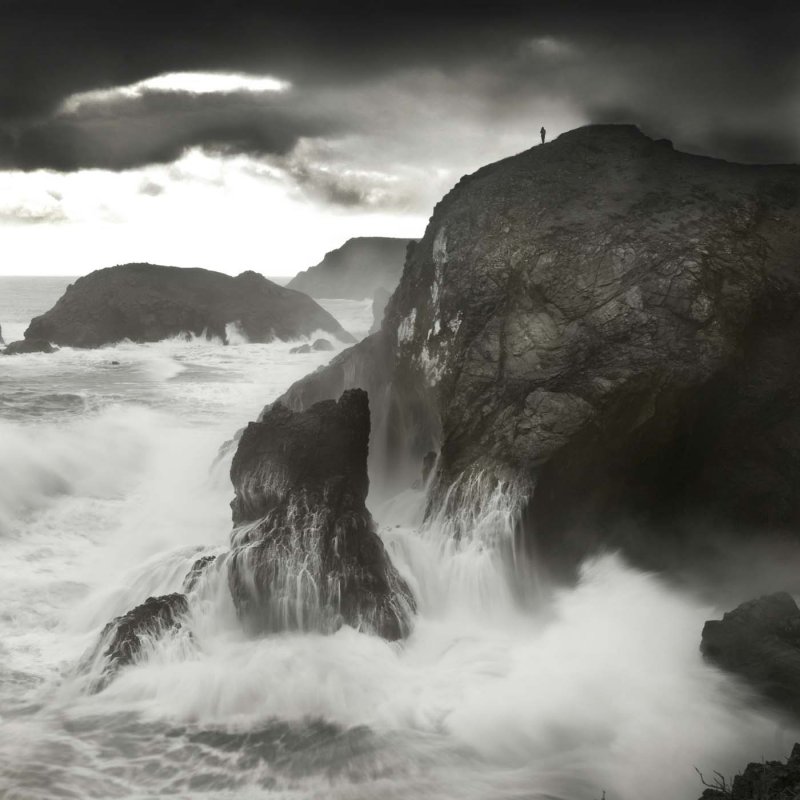 Dramatic grayscale image of sea crashing into cliff with a lone person standing at the top.