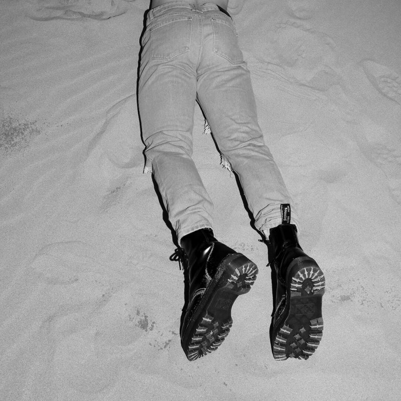 a man lying on his stomach in the sand, wearing light jeans and black boots 