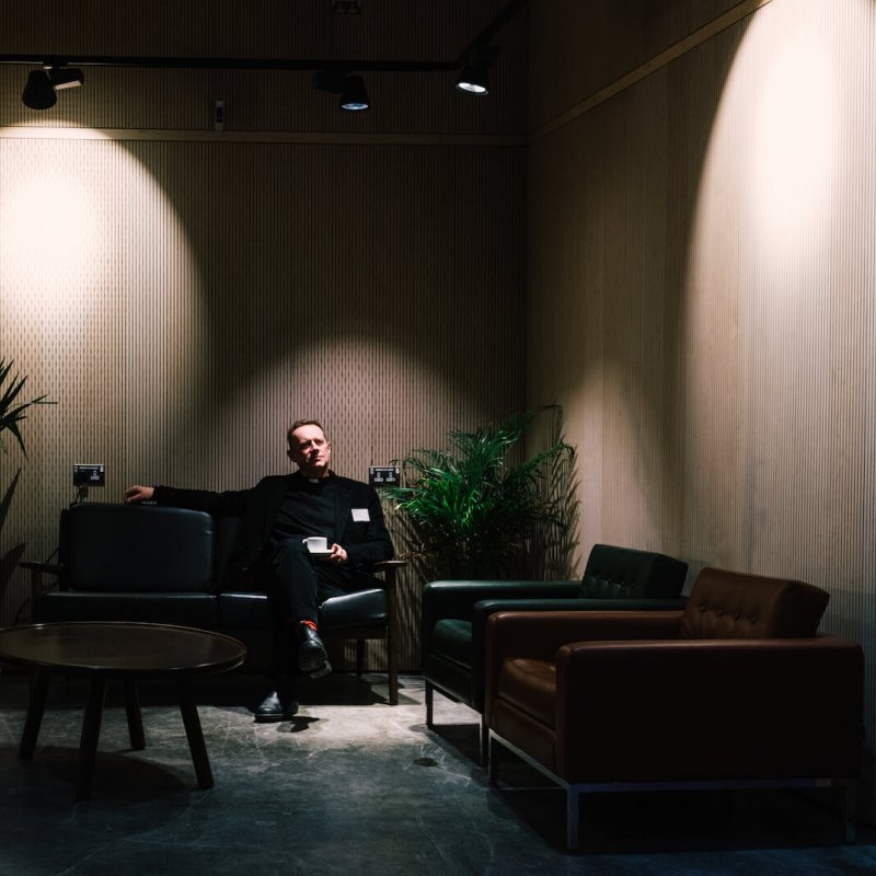 A man sat on a sofa in a darkly lit room with potted plants