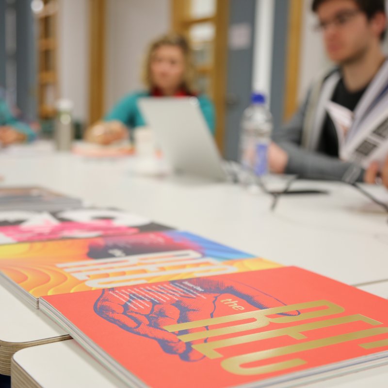 Design journals on a desk in the foreground, students working out of focus in the background.