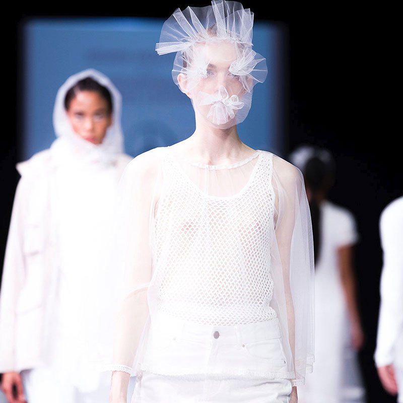 Models on a catwalk wearing white clothing and a netted face mask