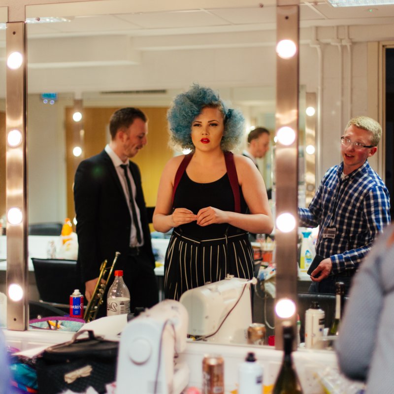 Backstage in the dressing room, actor with blue curly hair looking into mirror with lightbulbs.