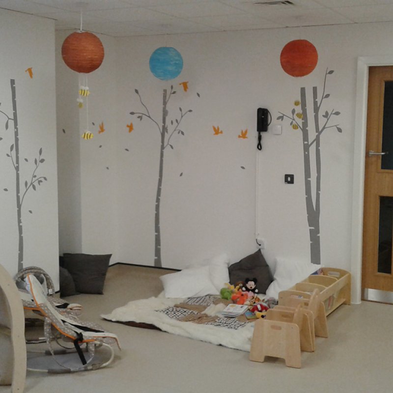 Room with children's toys, baby bouncers and tree illustrations on the wall.