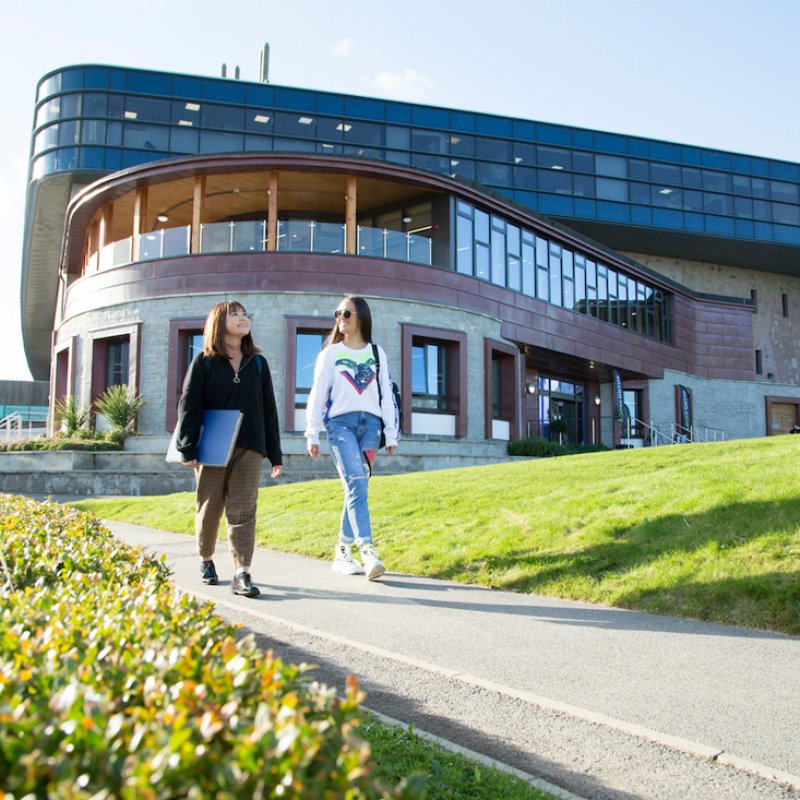 Two students walking past a large building on Penryn Campus