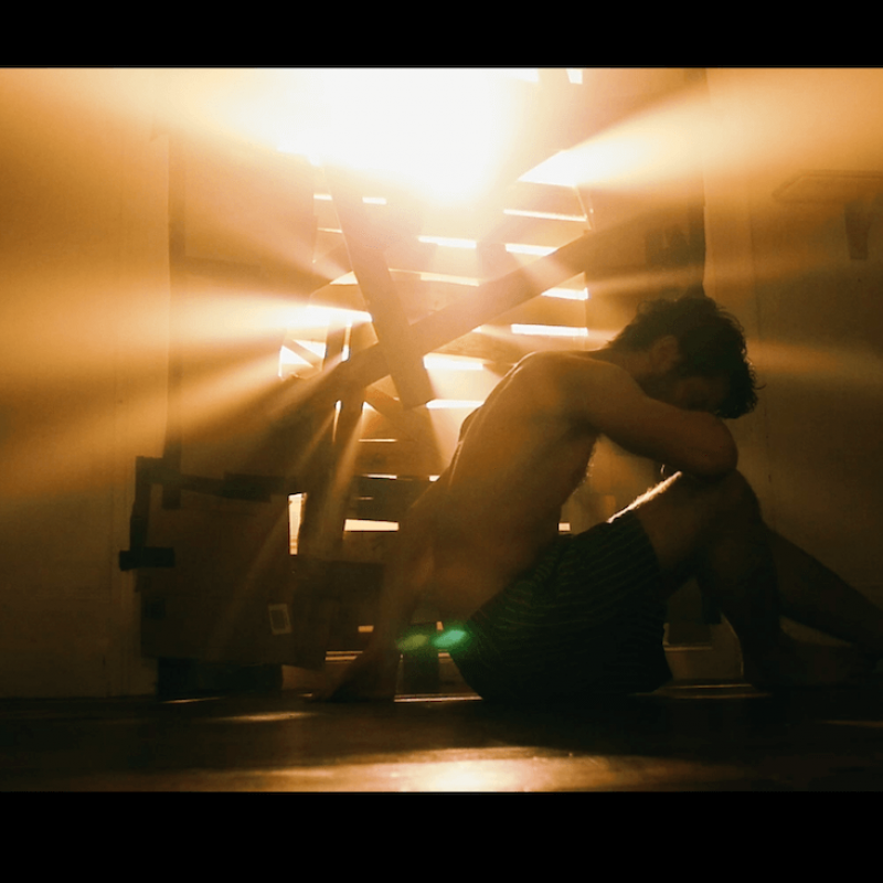 Film still of male in underwear with head in arms and light bursting through gaps in boarded up window.