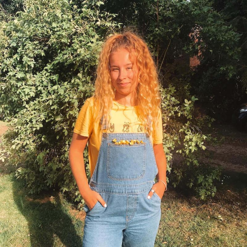 A Falmouth University student wearing denim dungarees and standing near bushes