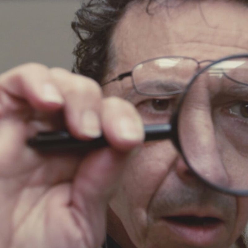 Film still, close up of a male face looking through a magnifying glass.