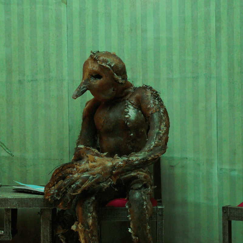 Strange creature sat on a desk in front of green striped wallpaper.