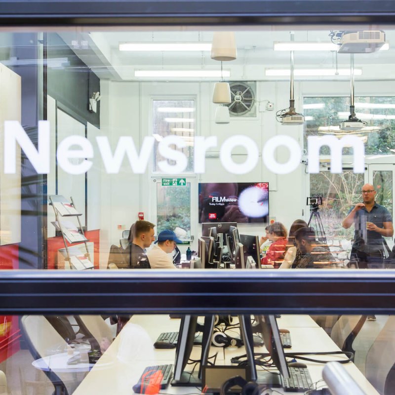 Through the window of the newsroom with lecturer talking to journalism students.