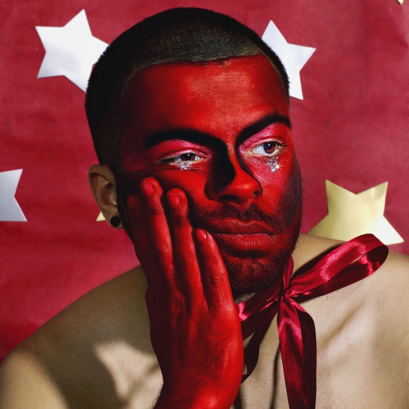 Man painted with red and tears of sparkles in front of red backdrop with stars.