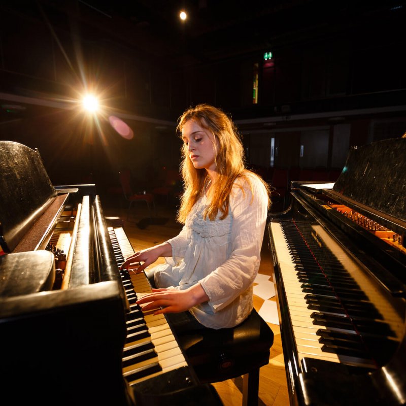 Female student sat between two pianos and playing one of them, sheet music on floor.