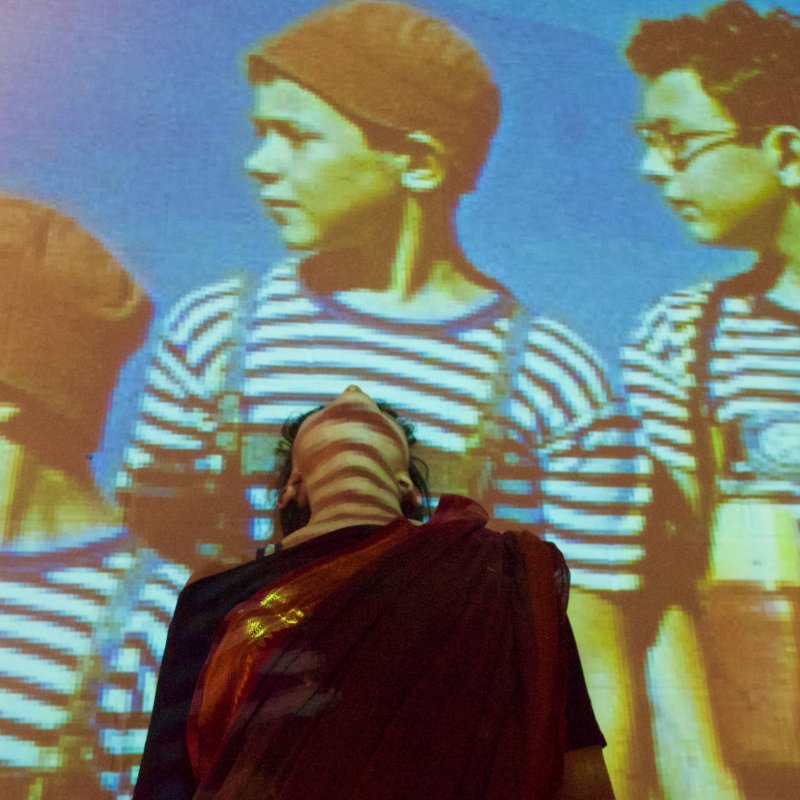 Woman leans backward towards projected image of boys in hats wearing stripy tops with dungarees 