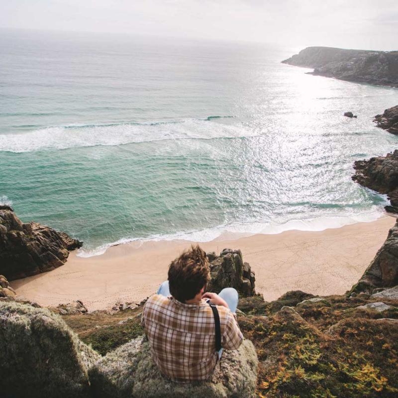 Man sat on the edge of cliff looking out to sun reflections on the water and coastline.