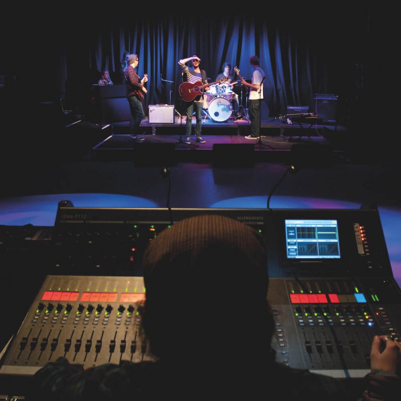 A student behind a sound desk with a view of a band performing
