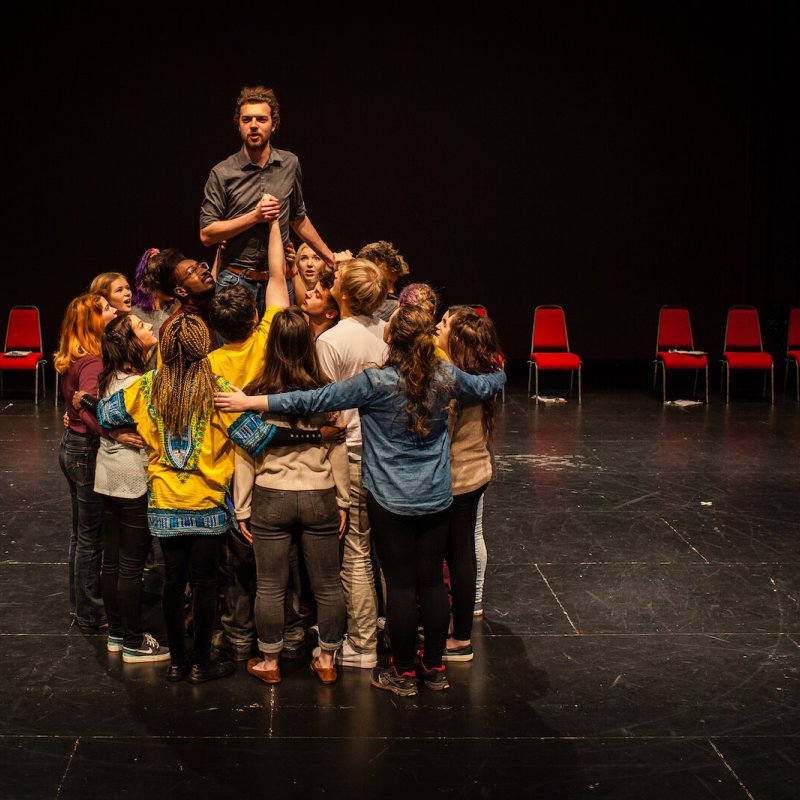 A group of acting students in a huddle with a male actor standing above them