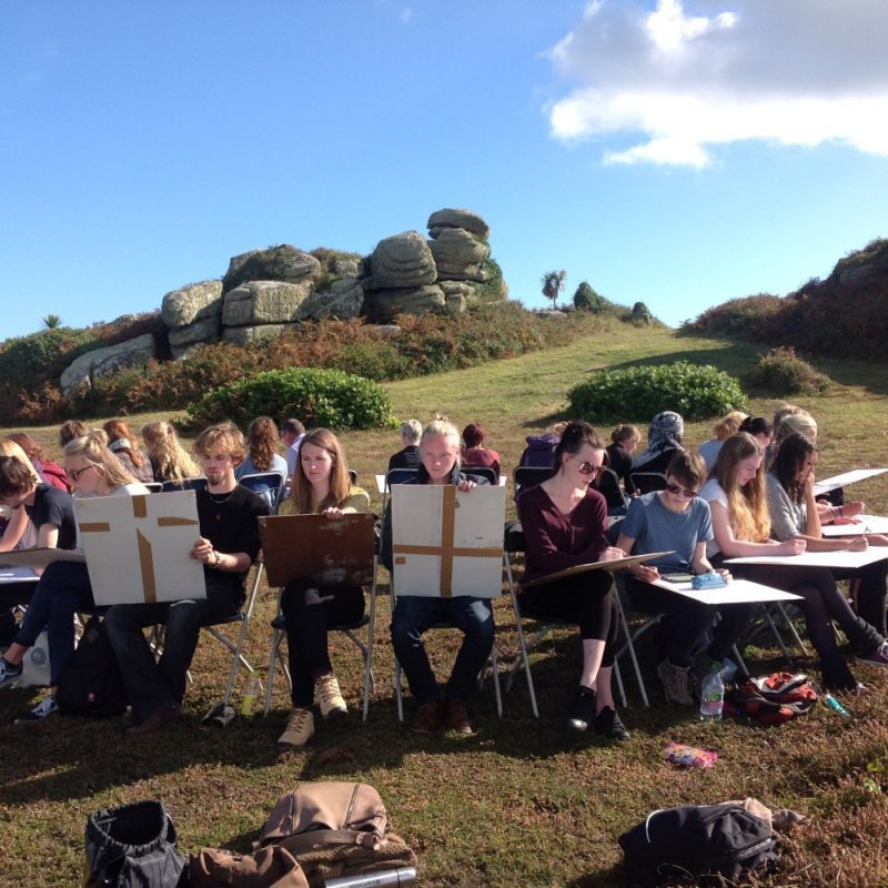 Group of students drawing outside sat on chairs and in rural landscape.