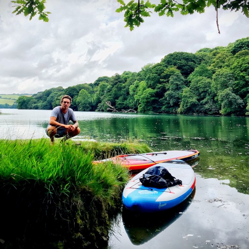 Falmouth University student crouching on the banks of the river with trees and paddleboards