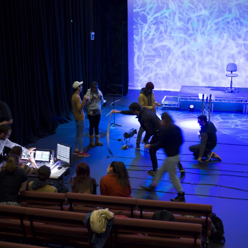 Group of students and teachers seated and moving around blue lit stage